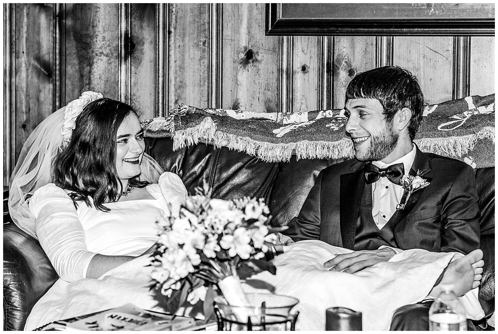 black and white portrait of bride and groom couple on wedding day looking at each other while seated on a leather couch