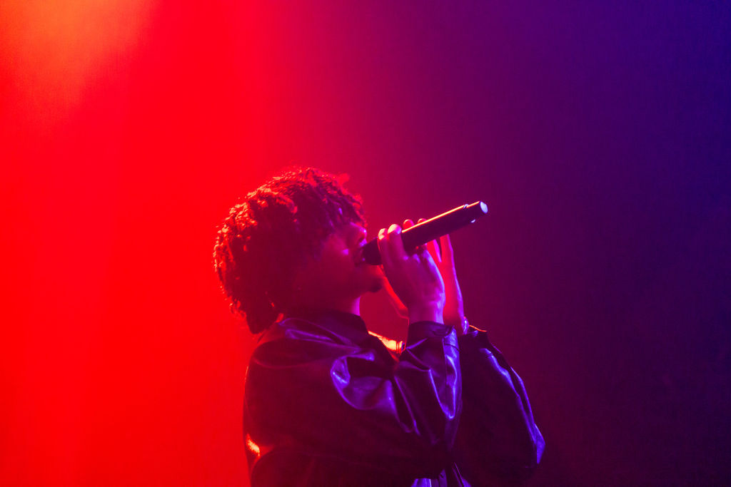 Atmospheric artist spotlight concert photo of 070 Shake raising a hand toward the crowd at the 9:30 Club in DC, surrounded by darkness and fractured light.