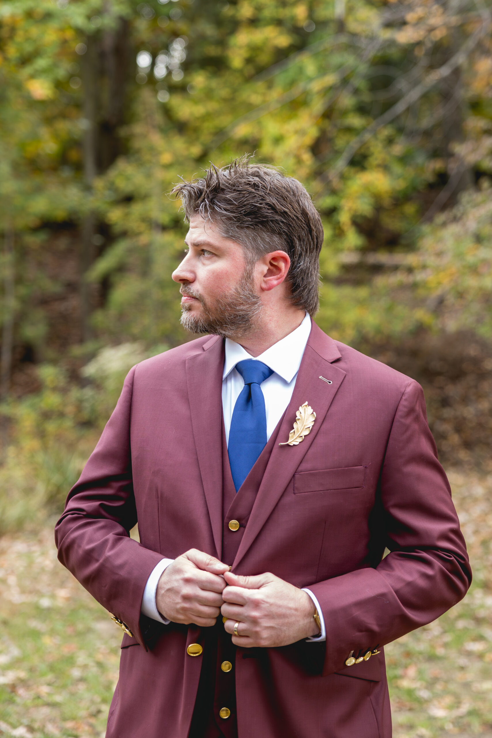 Groom portrait in maroon suit with blue tie and gold leaf boutonniere in fall woodland setting