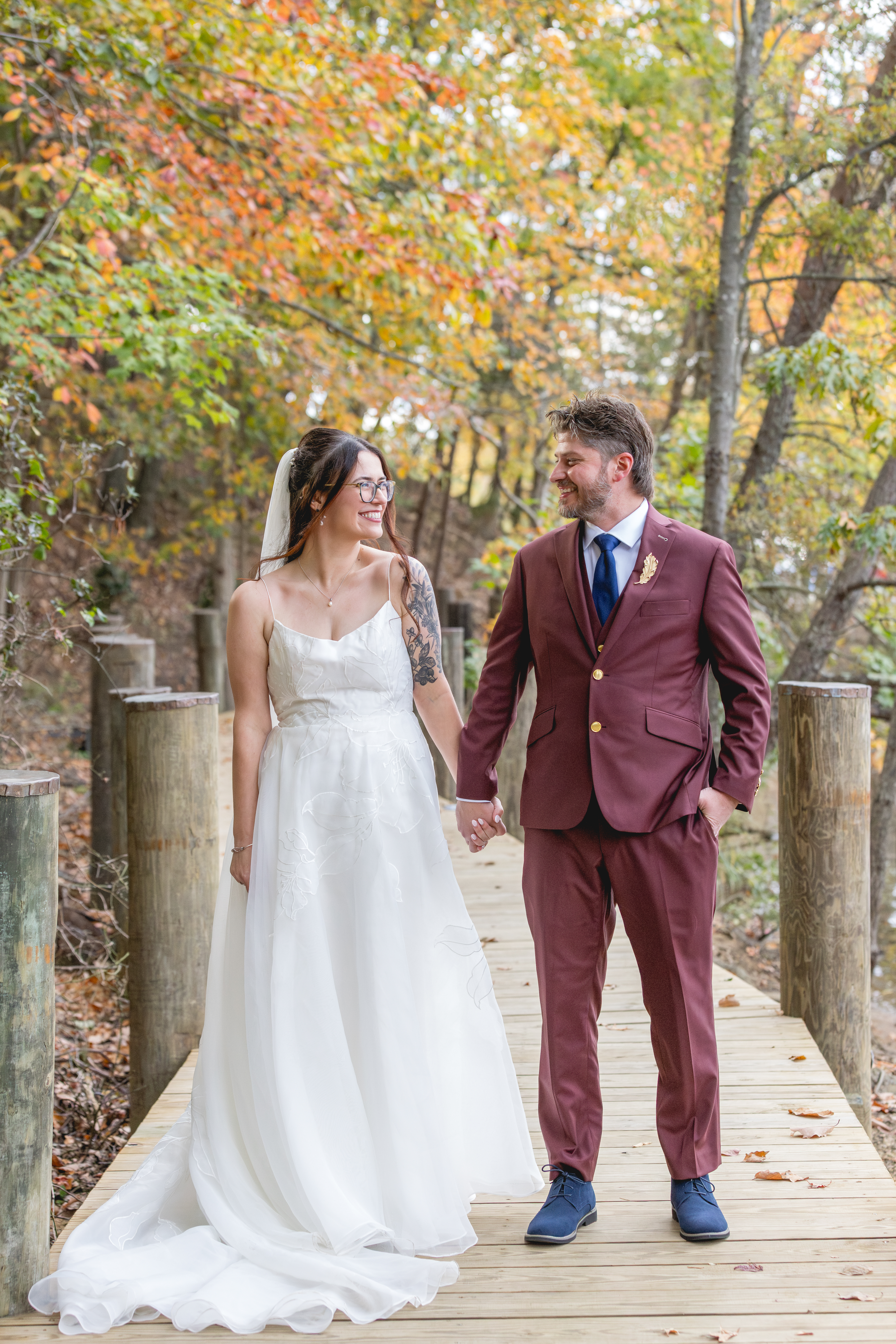 Bride and groom walking hand in hand along wooden dock with colorful fall leaves in Annapolis