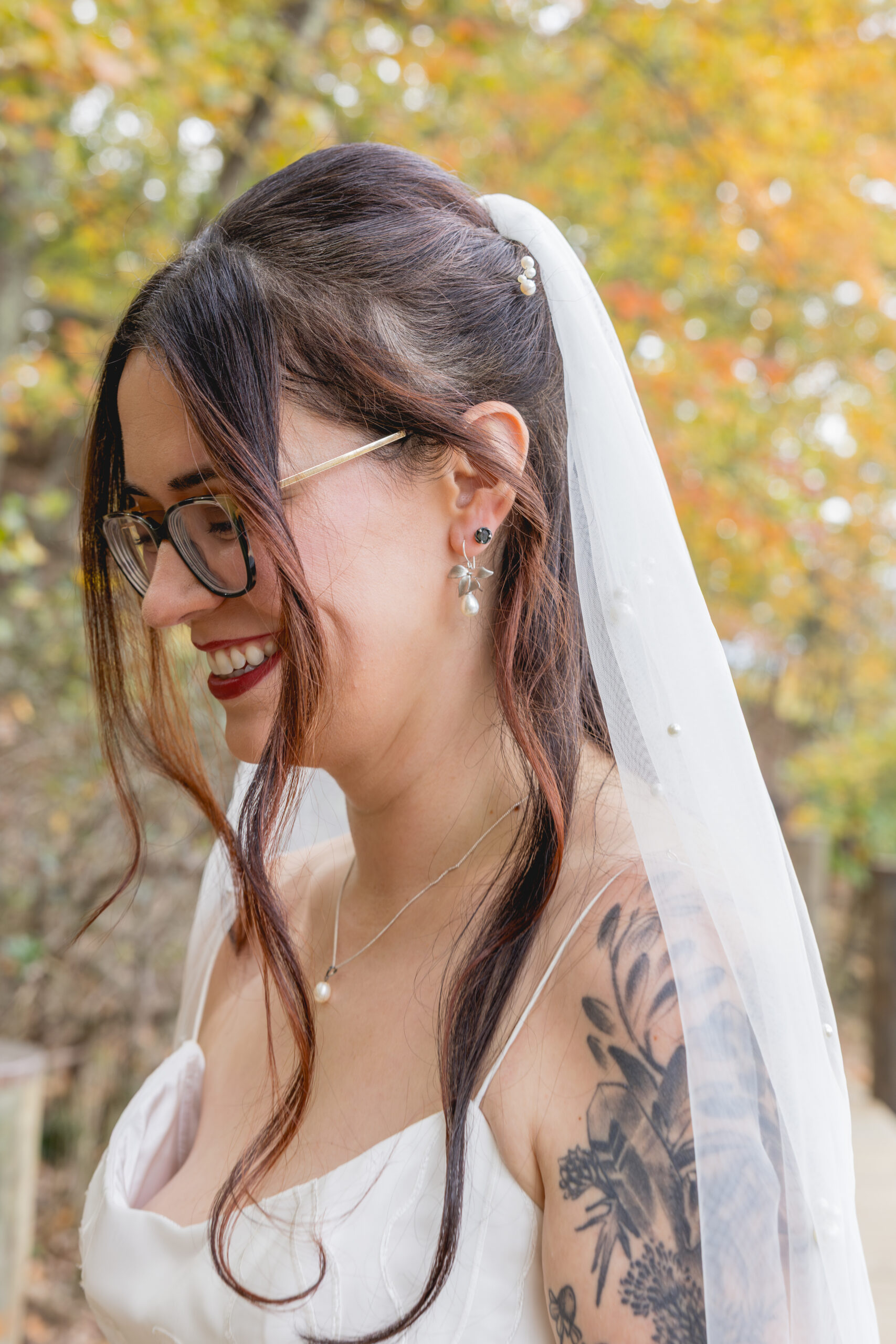Bride in veil and glasses looking down softly with autumn trees in background