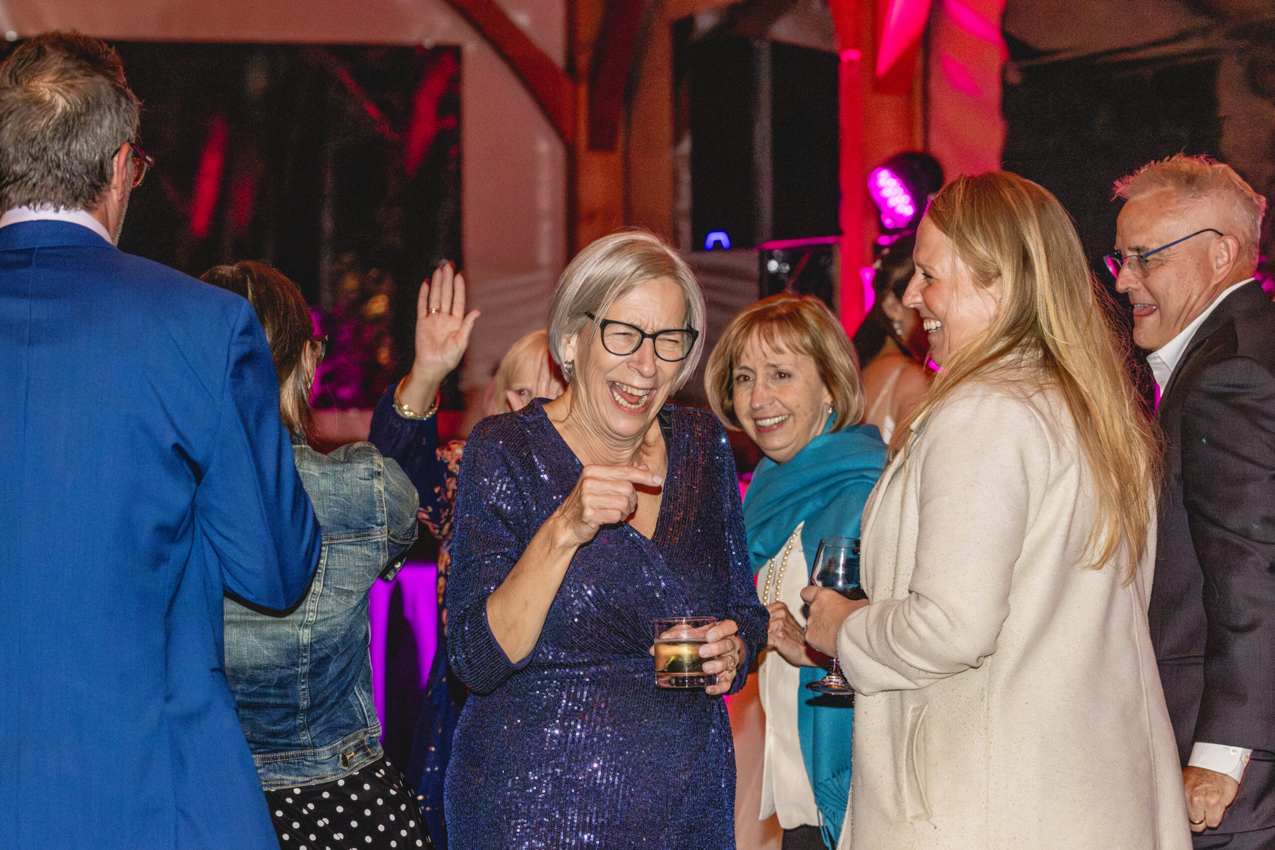 Wedding guest in blue sequin dress laughing and dancing while holding a drink under pink reception lighting