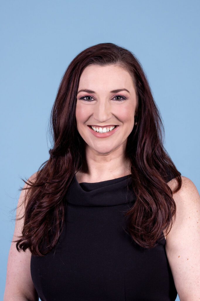 Professional headshot of a woman with dark auburn hair wearing a black sleeveless top, smiling on a blue background by Liz Stewart Photography DC