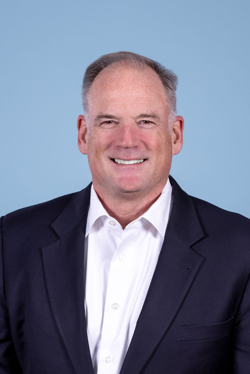 Corporate headshot of a man with gray hair wearing a navy blazer and open collar white shirt, smiling on a blue background by Liz Stewart Photography DC