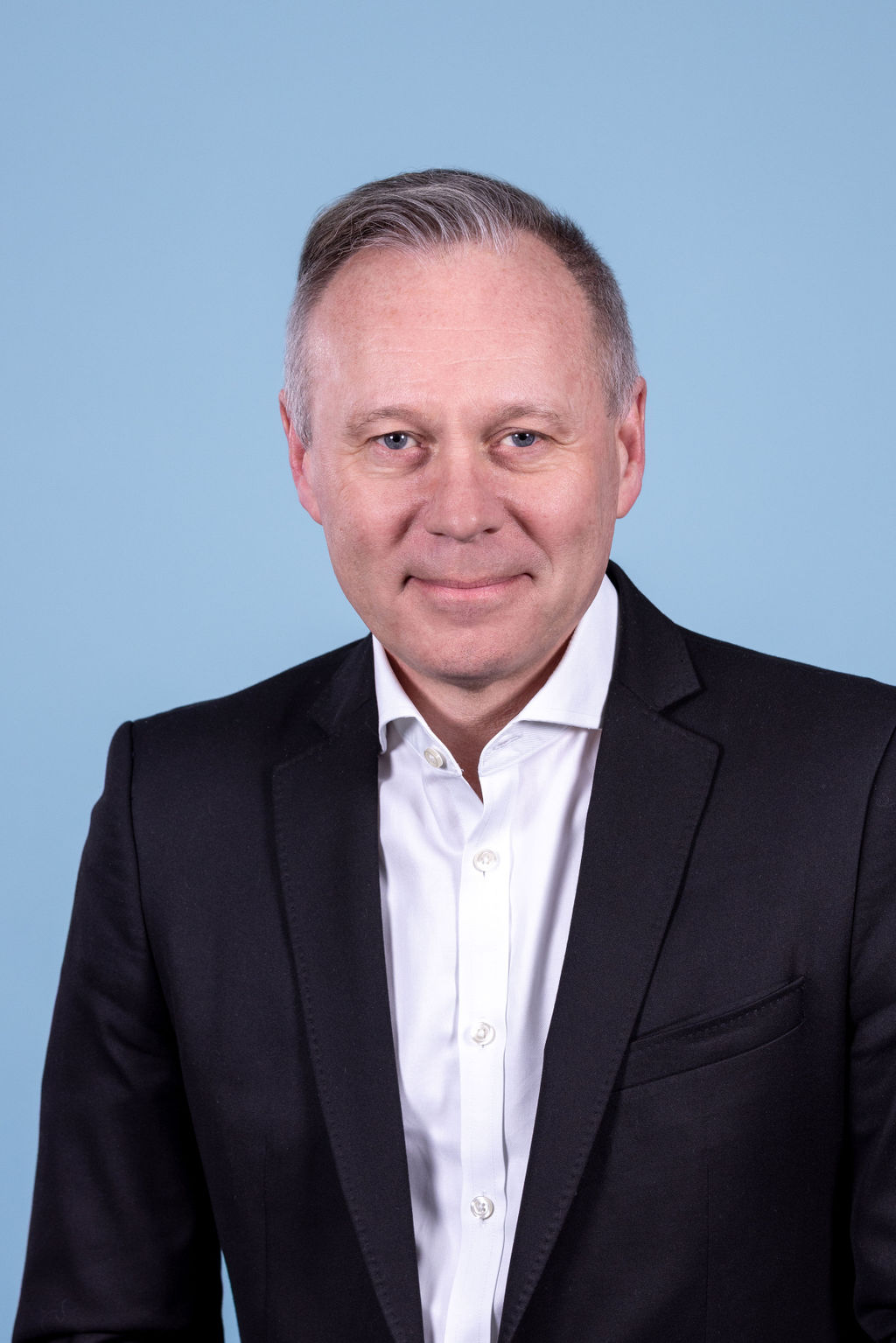 Professional headshot of a man with silver hair wearing a black suit and white shirt, photographed on a blue background by Liz Stewart Photography DC