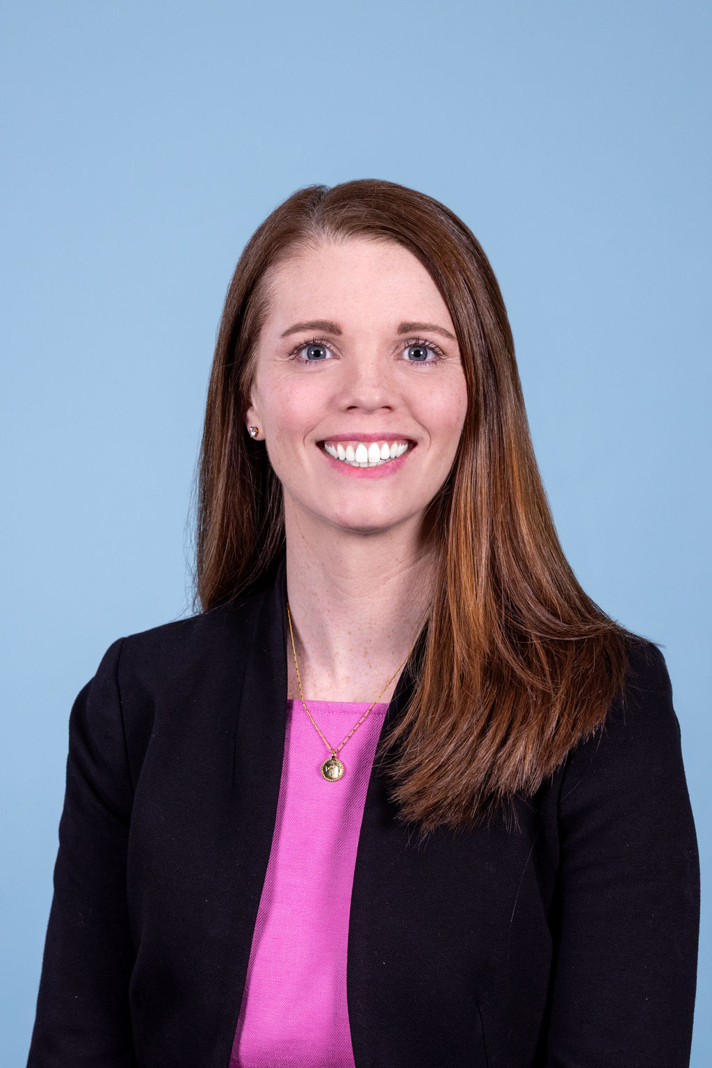 Corporate headshot of a woman with brown hair wearing a pink top and navy blazer, smiling warmly on a blue background by Liz Stewart Photography DC