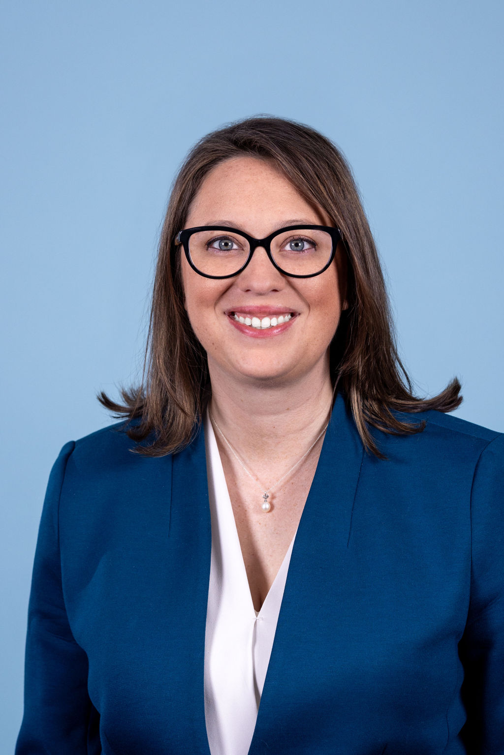 Professional headshot of a woman with brown hair and black frame glasses wearing a teal blue blazer, photographed on a blue background by Liz Stewart Photography DC