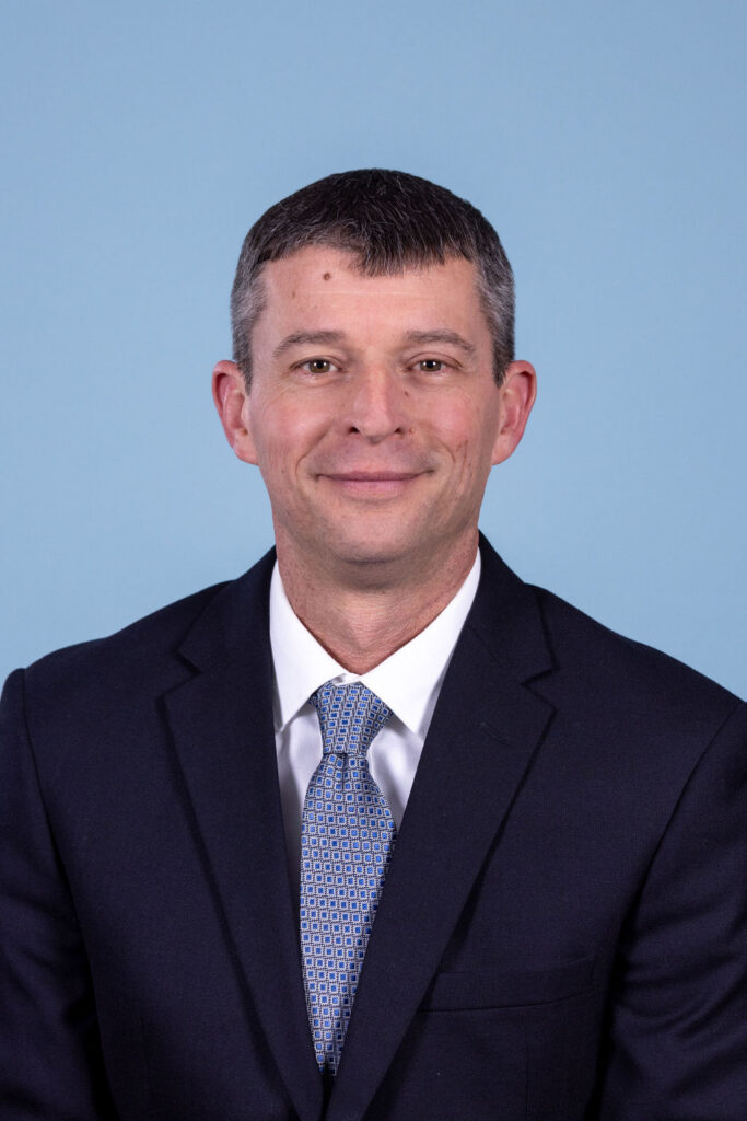 Professional headshot of a man with short dark hair wearing a navy suit and blue geometric tie, photographed on a blue background by Liz Stewart Photography DC
