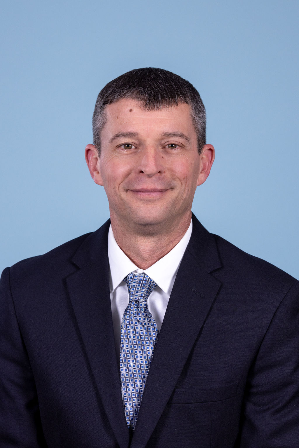 Professional headshot of a man with short dark hair wearing a navy suit and blue geometric tie, photographed on a blue background by Liz Stewart Photography DC