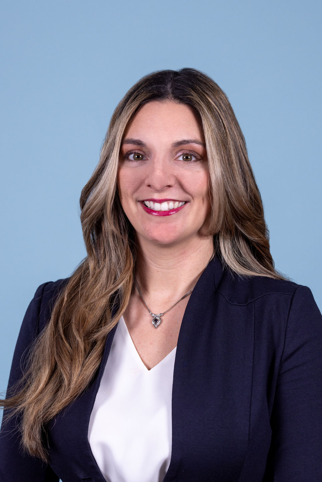 Corporate headshot of a woman with long highlighted brown hair wearing a navy blazer and white top, smiling on a blue background by Liz Stewart Photography DC