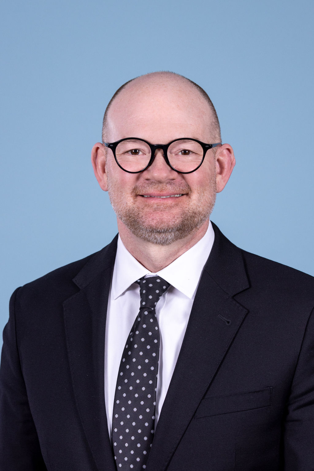 Professional headshot of a bald man with a beard wearing a black suit and polka dot tie, smiling on a blue background by Liz Stewart Photography DC
