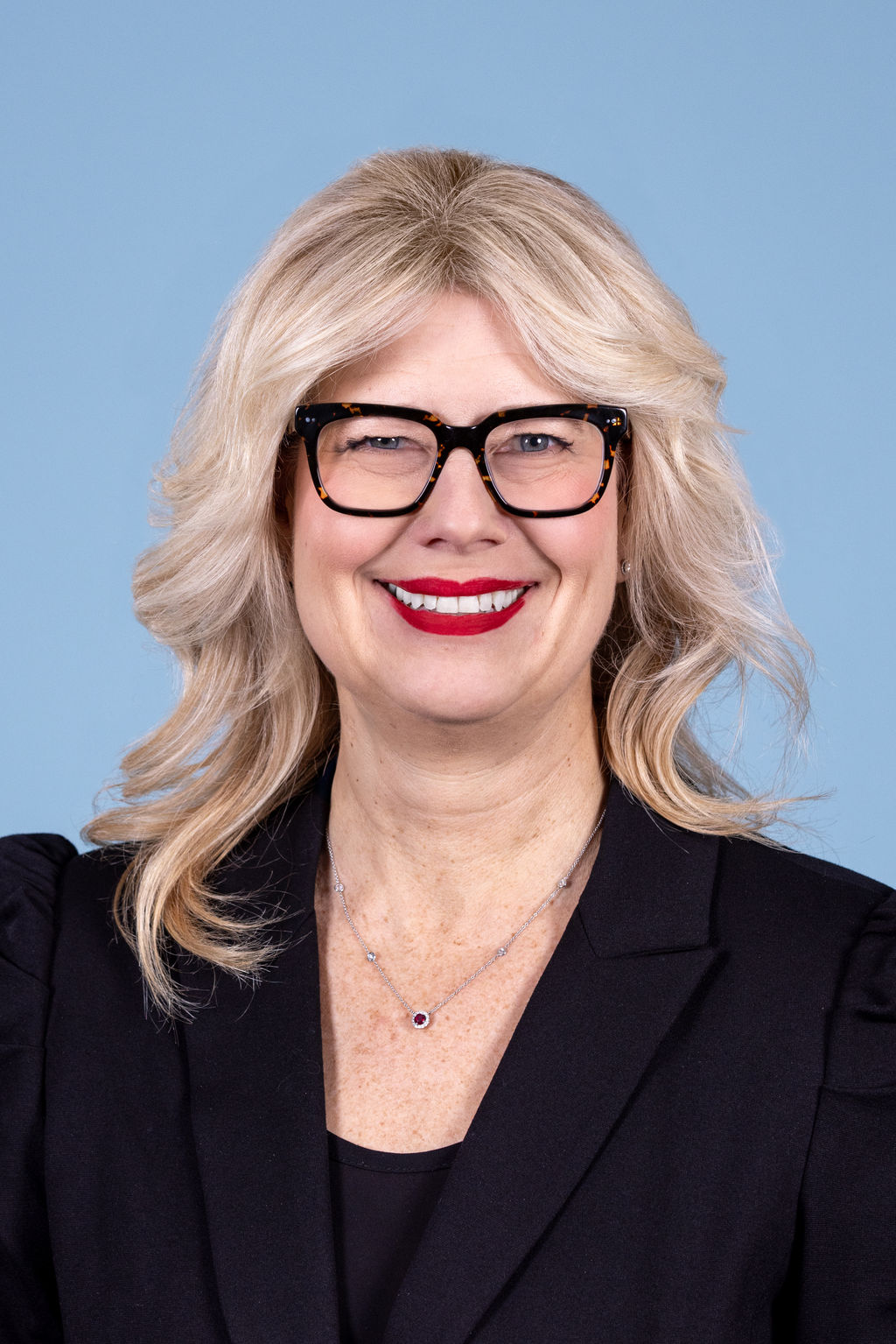Corporate headshot of a woman with blonde hair and tortoiseshell glasses wearing a black blazer, smiling with red lipstick on a blue background by Liz Stewart Photography DC