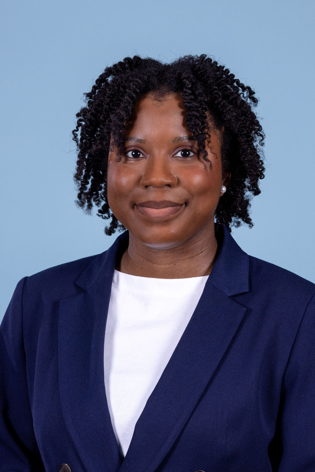 Professional headshot of a woman with natural curly hair wearing a navy blazer and white top, smiling on a blue background by Liz Stewart Photography DC