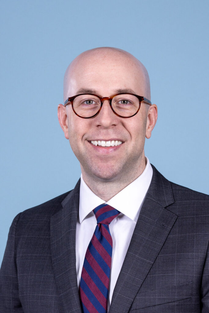 Professional headshot of a bald man wearing tortoiseshell glasses, a gray plaid suit, and a burgundy striped tie, photographed on a blue background by Liz Stewart Photography DC