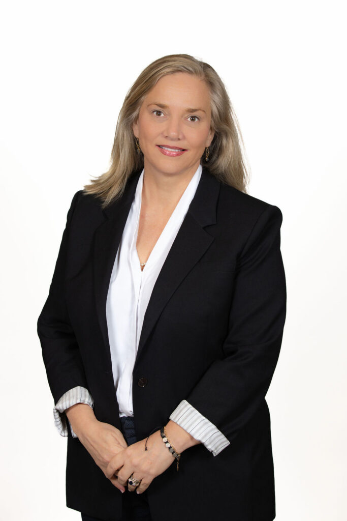 Corporate headshot of a woman with blonde and gray hair wearing a black blazer and white blouse, standing portrait on a white background by Liz Stewart Photography DC