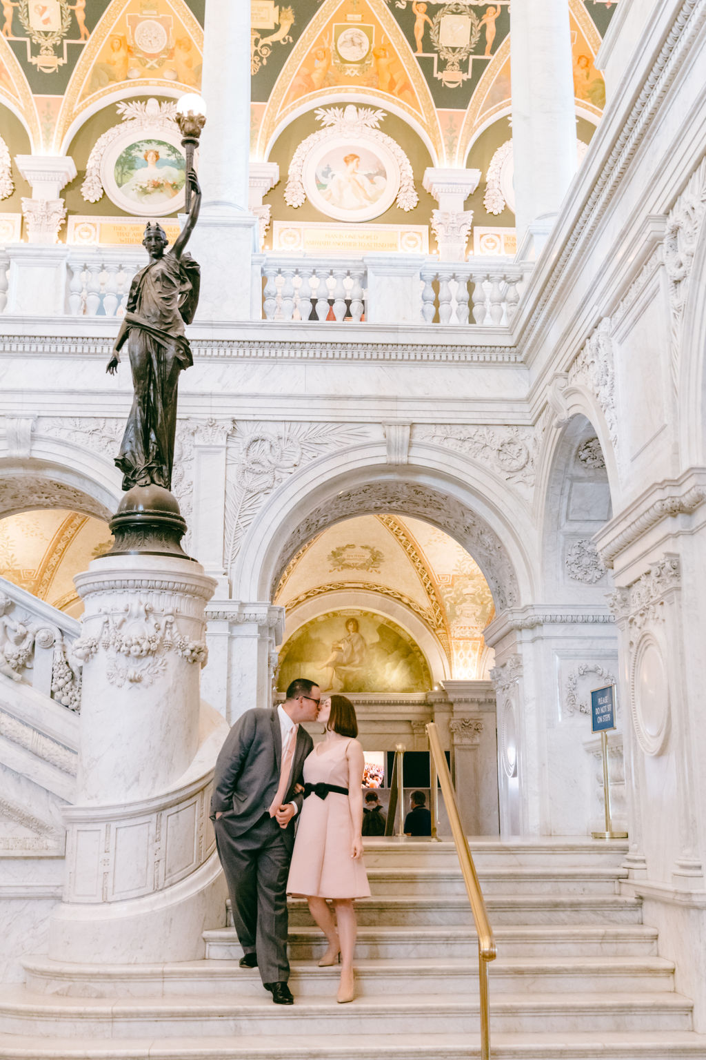 Engaged couple embracing on the upper staircase landing of the Library of Congress in Washington DC, framed by ornate columns and a painted gilded ceiling