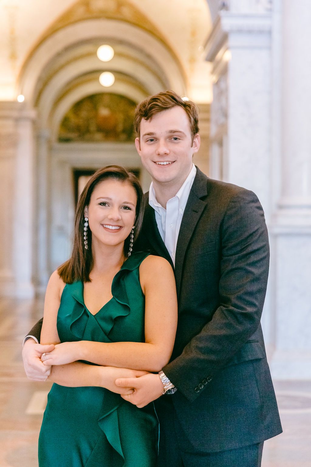 Engaged couple holding hands and leaning close together in a marble columned corridor of the Library of Congress in Washington DC, with painted golden arched ceilings overhead