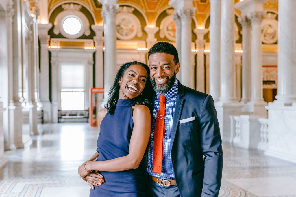 Candid portrait of a couple laughing together in the Great Hall of the Library of Congress, with golden painted arches and marble columns stretching behind them